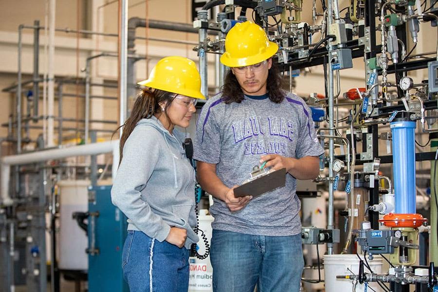 Two San Juan College students wearing hard hats checking a checklist in front of a series of pipes